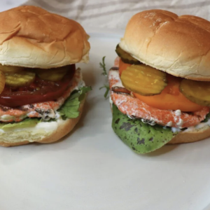 Two pieces of Salmon Burgers on a white plate packed with lettuce, tomatoes, and sauce.